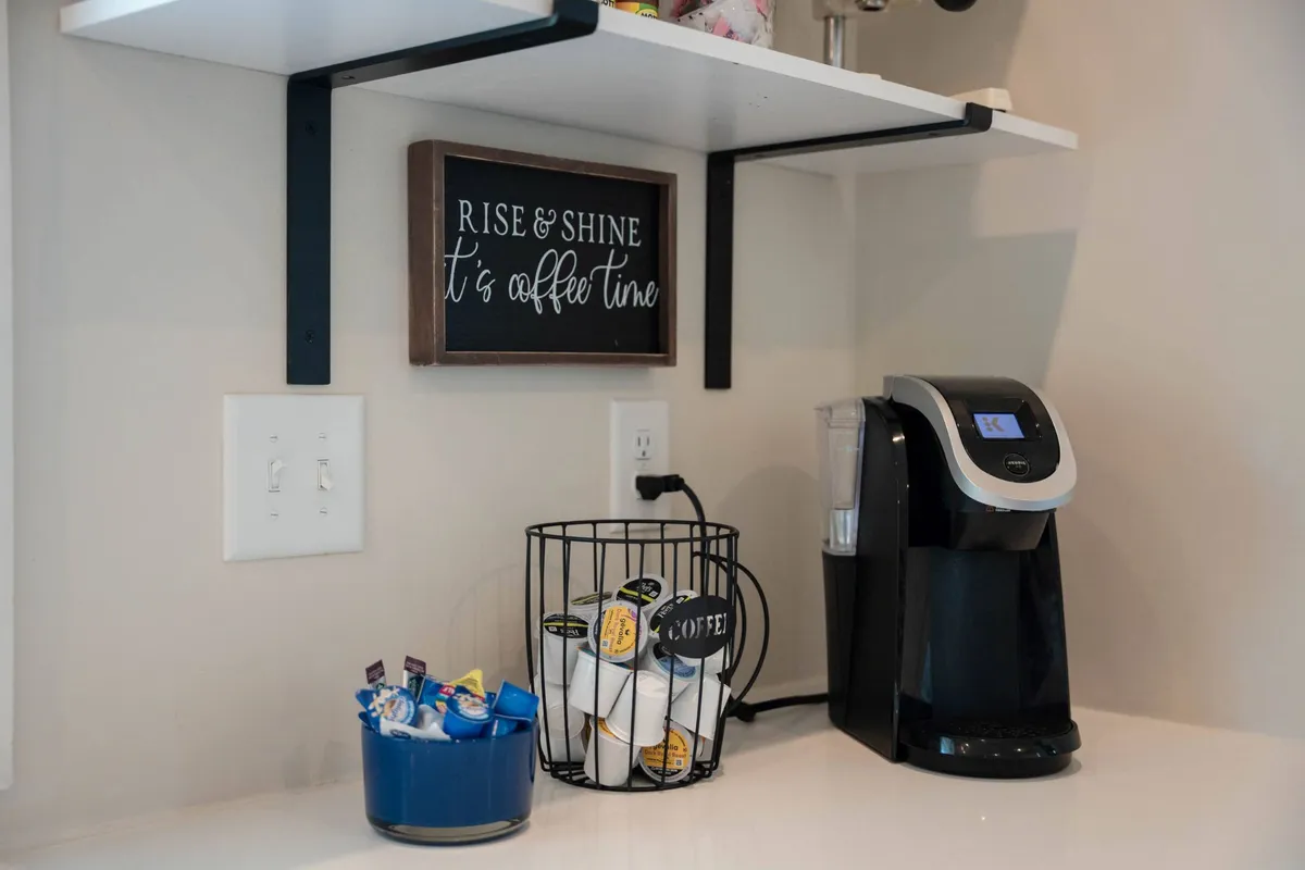 A close-up of the coffee station with a coffee maker, coffee pod holder, and a cheerful decorative sign, evoking the comfort of morning coffee routines and casual socializing.