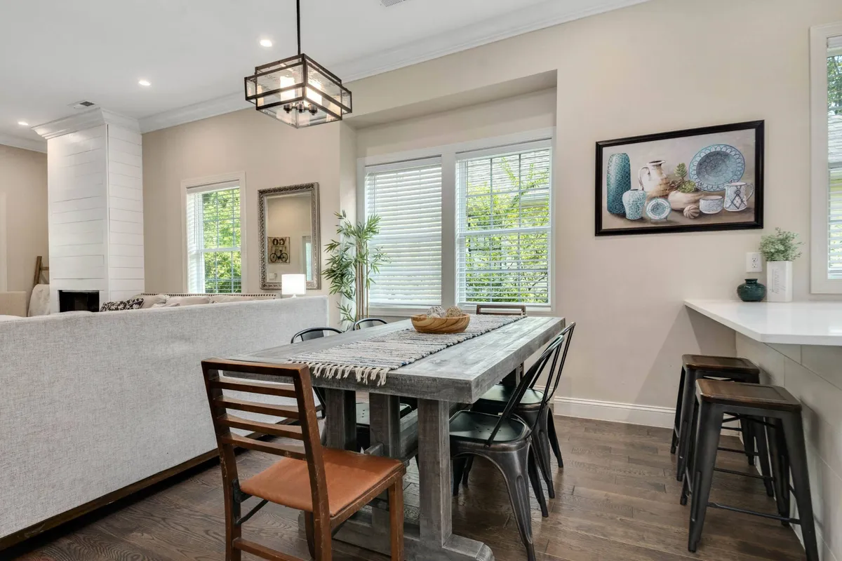 A cozy dining area featuring a wooden dining table set near windows that frame lush greenery outside, with tasteful artwork on the walls. The inviting atmosphere is ideal for family meals, friendly gatherings, or relaxed breakfasts.