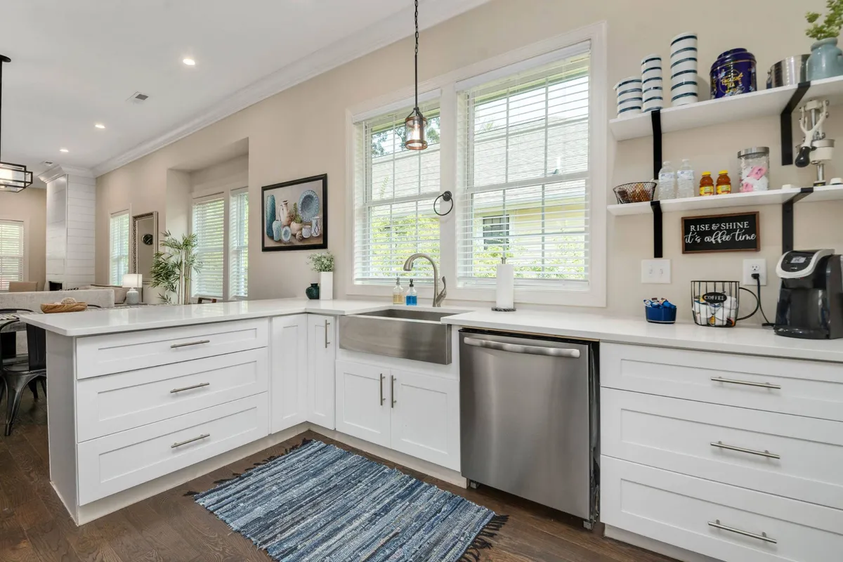 A bright, cheerful kitchen featuring a central island with a sink, stylish shelving with decorative accents, and a large window allowing natural light to fill the space. The layout invites group cooking and socializing, with ample countertop space for prepping meals.
