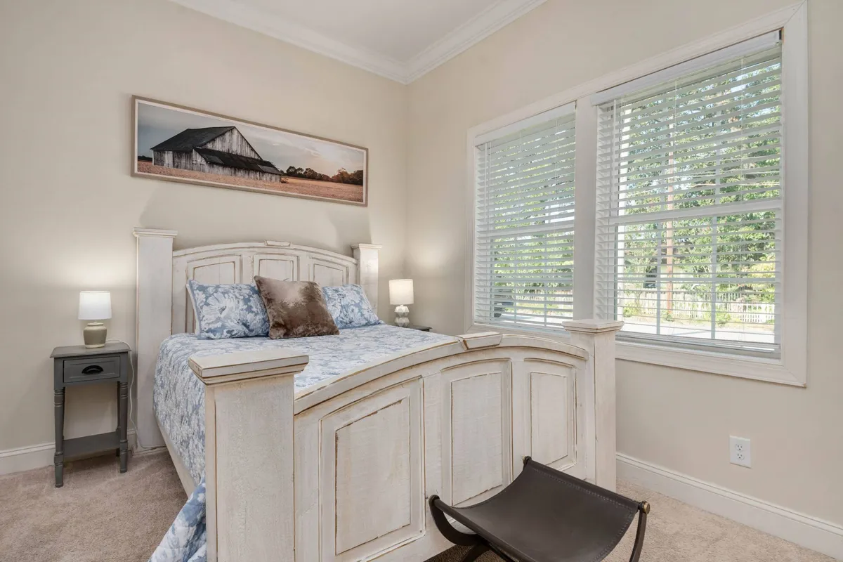 A peaceful queen guest room with a comfortable bed topped with patterned bedding, framed wall art, a nightstand with a lamp, and a window shaded by blinds. The space feels restful and cozy, ideal for guests seeking a quiet place to recharge.