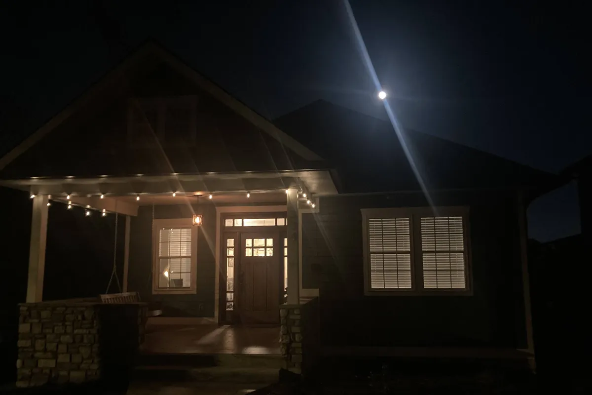 The image shows the exterior front porch of the property illuminated by the warm glow of lighted windows under the soft moonlight. Cozy seating is visible, inviting guests to relax and socialize outdoors as the evening sets in.