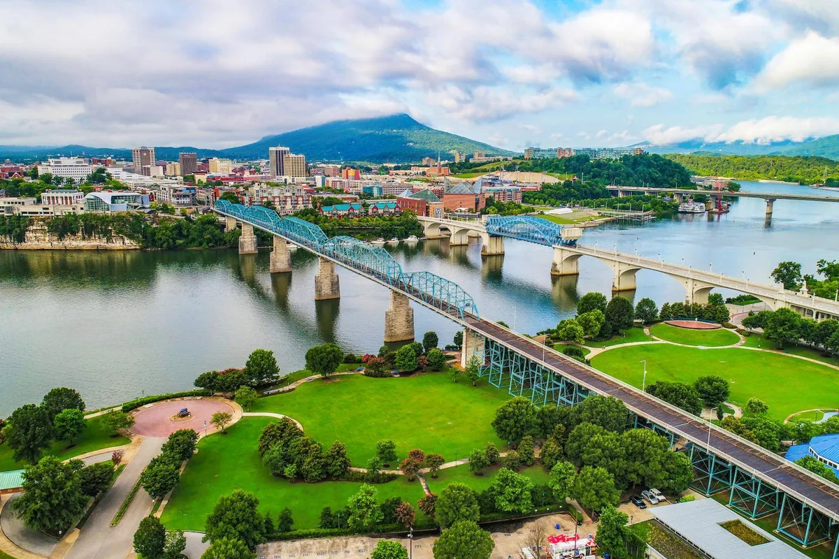 A sweeping view over the river with bridges, city skyline in the distance, lush green parks, and walking paths. The scene captures Chattanooga's vibrant riverfront and the proximity to outdoor recreational areas and attractions.