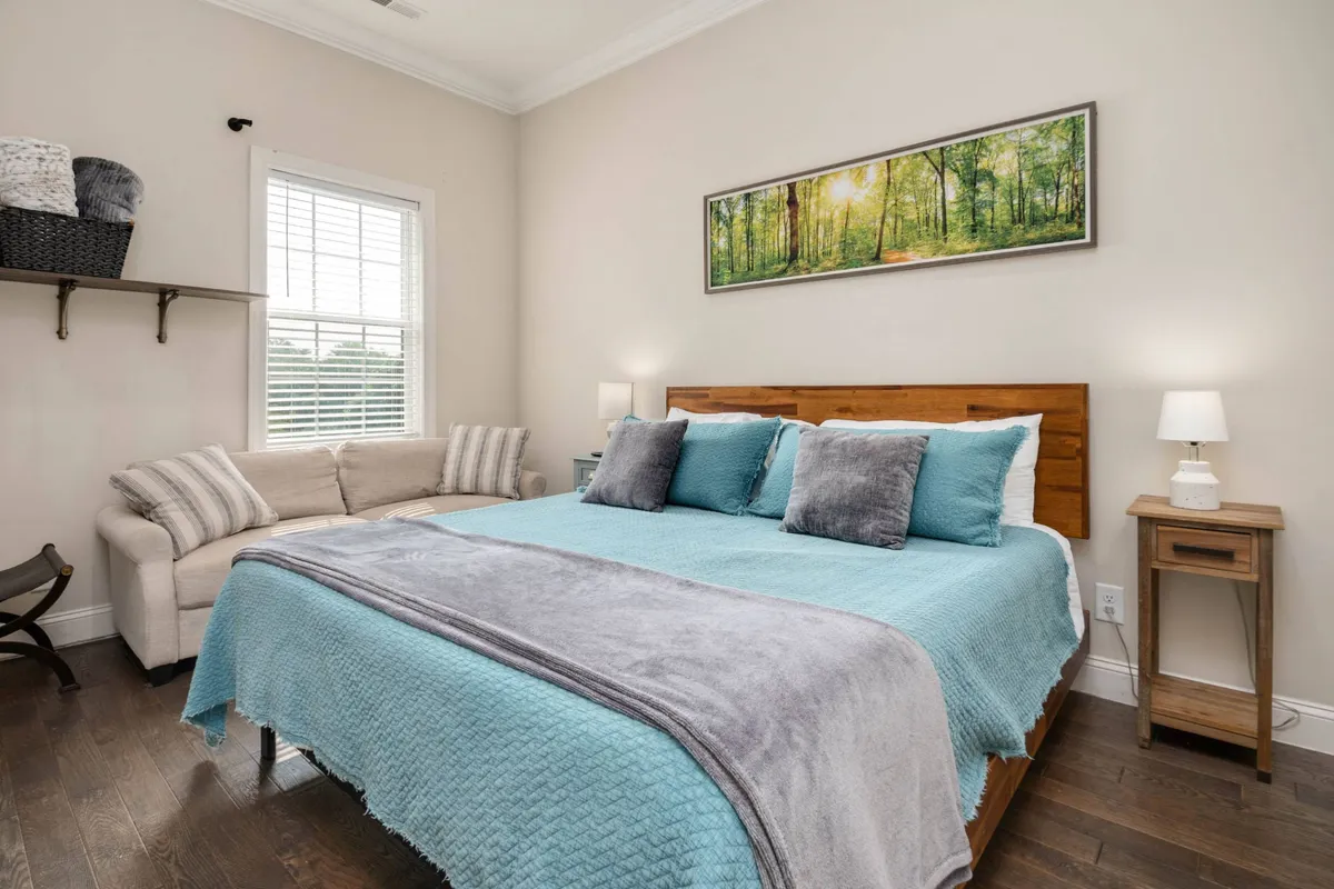 The image features a serene bedroom with a plush bed dressed in turquoise bedding, striking artwork above the headboard, and a cozy seating area with a sofa perfect for private relaxation. Natural light softens the space, creating an inviting atmosphere ideal for winding down after a busy day.