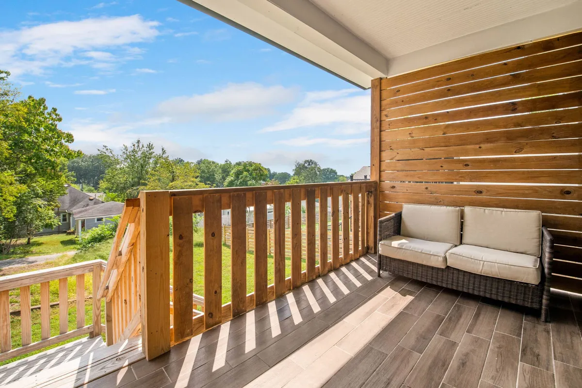 This image shows a wooden deck with a comfortable seating area and a wooden railing, overlooking a lush green landscape under a blue sky. The arrangement invites relaxation and morning rituals, with ample space for sipping coffee and enjoying the tranquil view.