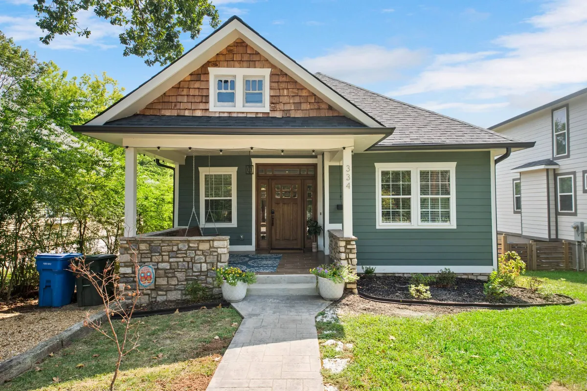 The image showcases the inviting exterior of The Swallowtail, a charming single-family bungalow. A welcoming front porch adorned with flower pots, a distinctive wooden door, elegant stonework, and a classic shingle roof are all visible. The scene hints at spaces perfect for family gatherings, morning coffee, and children playing in the yard, set within a quiet, historic Chattanooga neighborhood.