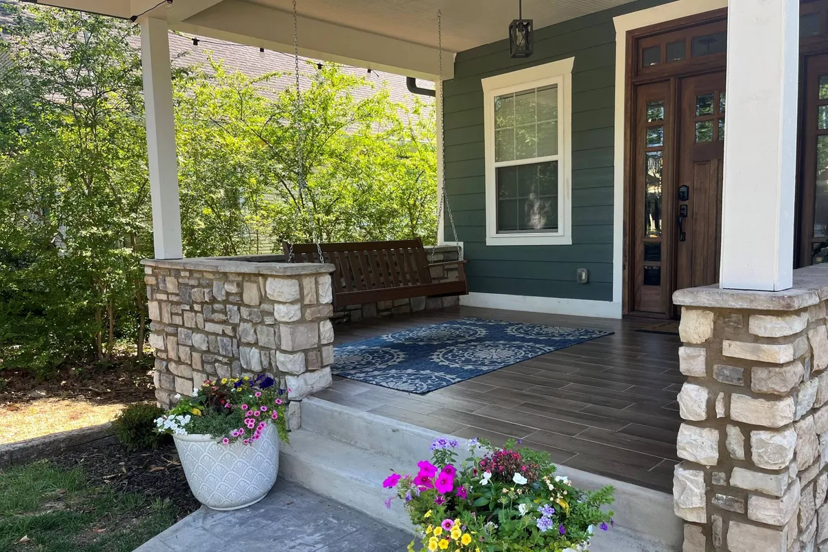 An inviting exterior shot of a swing bench on a porch flanked by stone pillars and decorated with flower pots and a cozy rug. The setting is surrounded by greenery and has a relaxing, outdoor ambiance—suggesting the home’s connection to its outdoor yard.