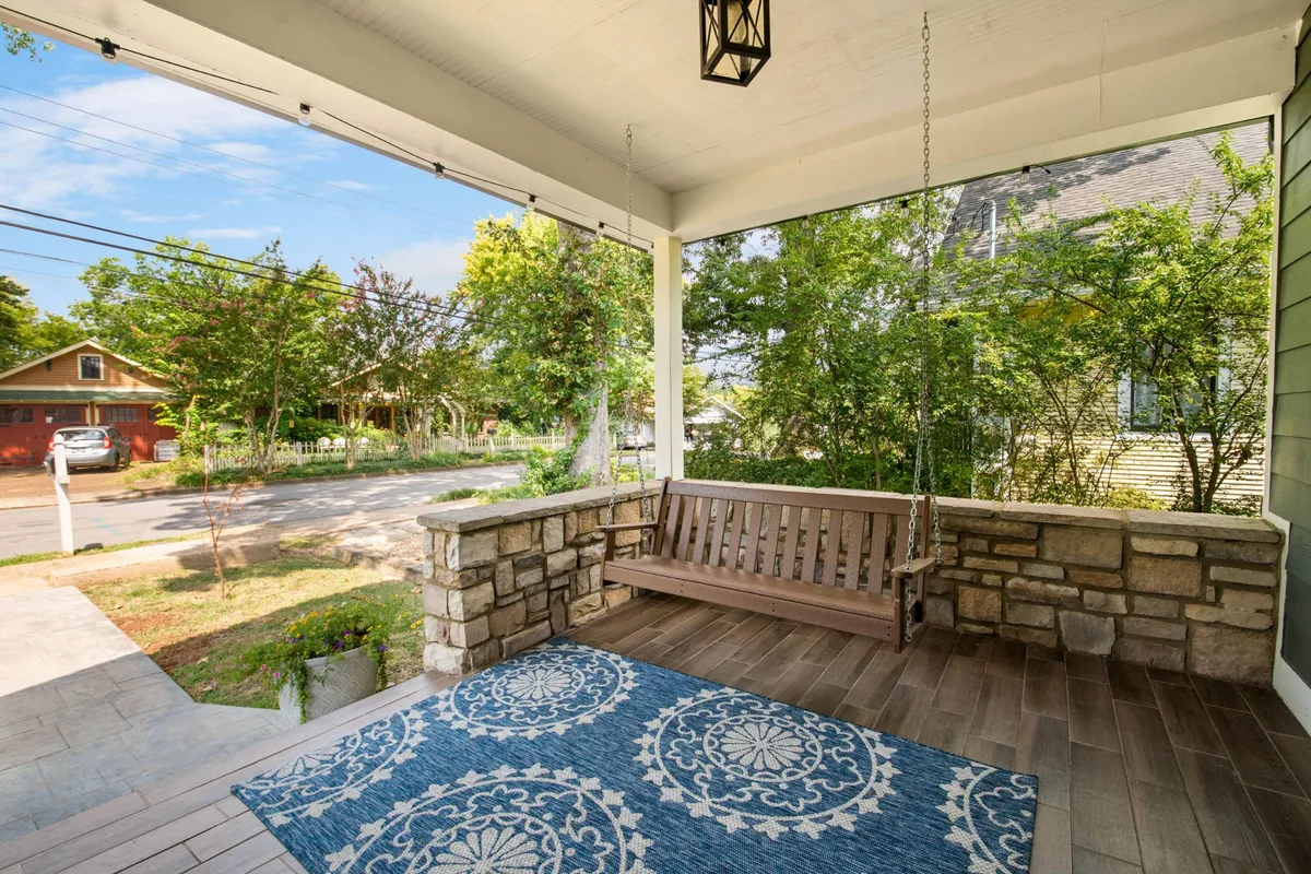 A cozy swinging bench sits on a porch, paired with a decorative rug and set against a stone wall surrounded by greenery. The porch exudes tranquility and invites moments of relaxation, reading, or watching the sunset.