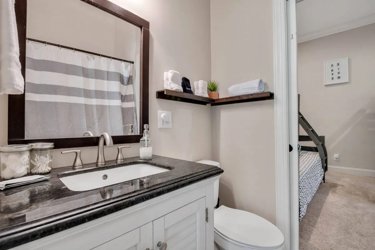 An inviting bathroom scene featuring a modern sink with faucet, a large mirror, neatly stacked towels on a shelf, a striped shower curtain, and a decorative plant. The space is bright, fresh, and fully stocked for guest comfort.